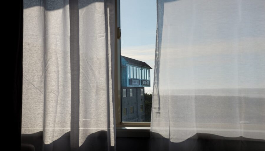 A view through a window with sheer white curtains on either side reveals a building in the distance with a large sign on its facade. The sky is clear and blue, and the surrounding area outside the window appears bright and sunny.