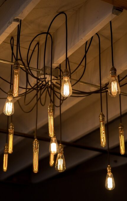 A group of vintage-style Edison light bulbs is suspended from a ceiling, hanging at various lengths. The bulbs are lit, casting a warm glow in the dimly lit room. Exposed wooden beams and wiring are visible above the lights.