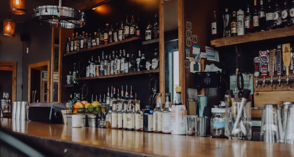 A dimly lit bar with a fully stocked shelf of liquor bottles behind the counter. Bar tools, including cocktail shakers and glasses, are on the counter. Hanging lamps with warm lighting illuminate the setting.