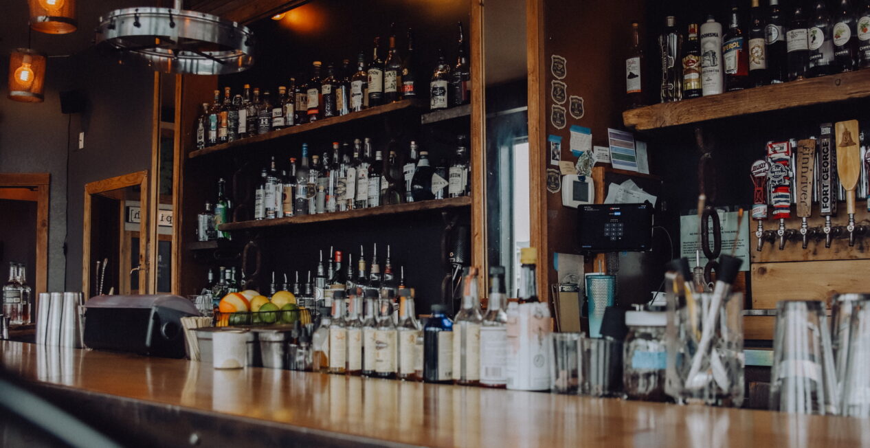 A dimly lit bar with a fully stocked shelf of liquor bottles behind the counter. Bar tools, including cocktail shakers and glasses, are on the counter. Hanging lamps with warm lighting illuminate the setting.