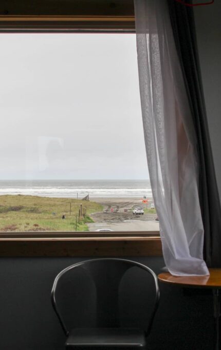 A view of the ocean and grassy dunes is seen through a window with light gray curtains from inside a dark room. Near the window, there is a small round table with a chair beside it.