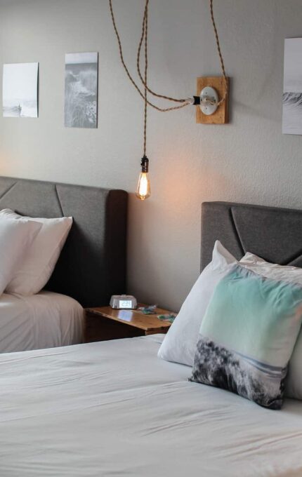 A bedroom with two beds featuring white linens, gray upholstered headboards, and decorative pillows. Pendant lights hang above the nightstands, while three framed pictures of waves decorate the wall. A window allows natural light to enter the room.