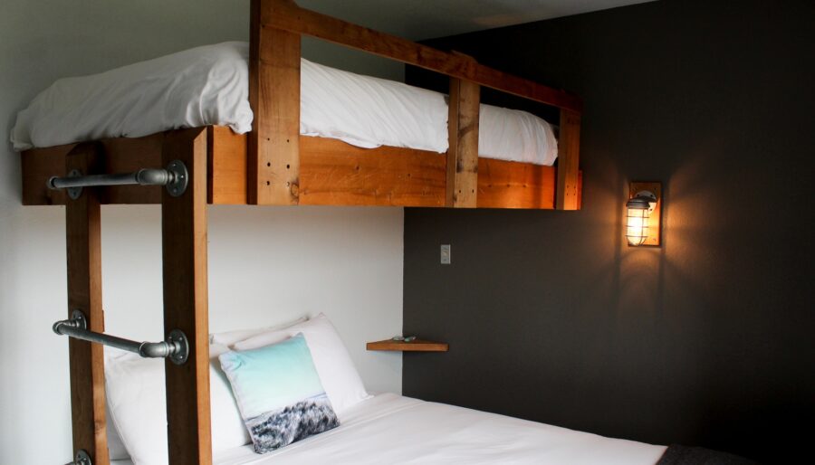 A room with a bunk bed setup. The top bunk features a wooden frame with white bedding. The bottom bed, also with white bedding, includes a pillow with a landscape design. The wall behind is gray with a mounted industrial-style light fixture.