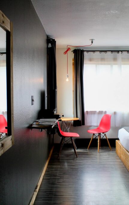 A modern hotel room with two twin beds, a small wooden desk with a laptop, and two bright red chairs. A large mirror is mounted on the dark wall. The room has large windows with white curtains, letting in natural light. The floor is dark wood.