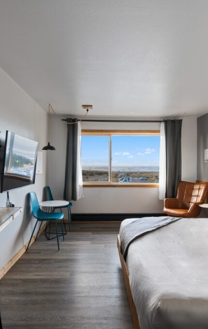 A minimalist hotel room with a large bed, a wooden desk with a lamp, two chairs, a wall-mounted TV, and a window. The room has a modern decor with wood accents and photographs on the wall. Outside the window, a landscape view is visible.