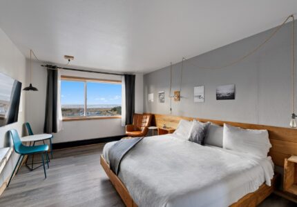 A neatly arranged modern bedroom features a large bed with white linens, wooden headboard, and side tables. The room has a large window with a scenic view, a small round table with two blue chairs, and a brown leather chair in one corner.