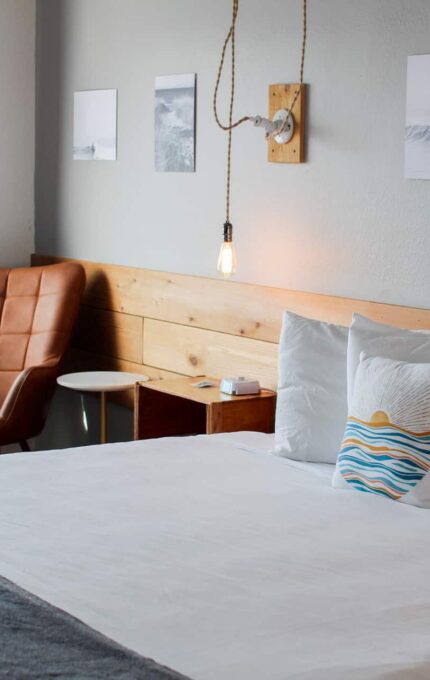 A neatly made bed with white linens and a gray blanket is in a modern room. An upholstered chair and a small circular table are placed next to the bed. Three framed pictures are hung on the wall, and an exposed light bulb hangs above the bed.