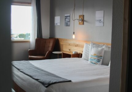 A modern bedroom with a large bed featuring white linens and a gray throw. There is a brown cushioned chair near a window with light curtains. Three small framed pictures are on the wall above the bed, and a single exposed light bulb hangs on the wall.