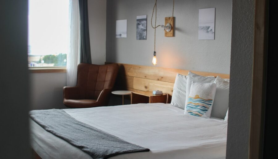 A modern bedroom with a large bed featuring white linens and a gray throw. There is a brown cushioned chair near a window with light curtains. Three small framed pictures are on the wall above the bed, and a single exposed light bulb hangs on the wall.