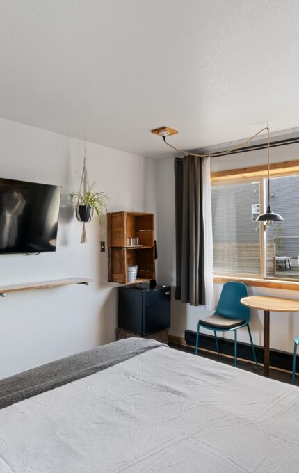 A cozy, modern bedroom features a wall-mounted TV, a small table with two chairs by a window with gray curtains, a bed partially visible in the foreground, and decor including a hanging plant, a framed picture, and a wooden shelf.