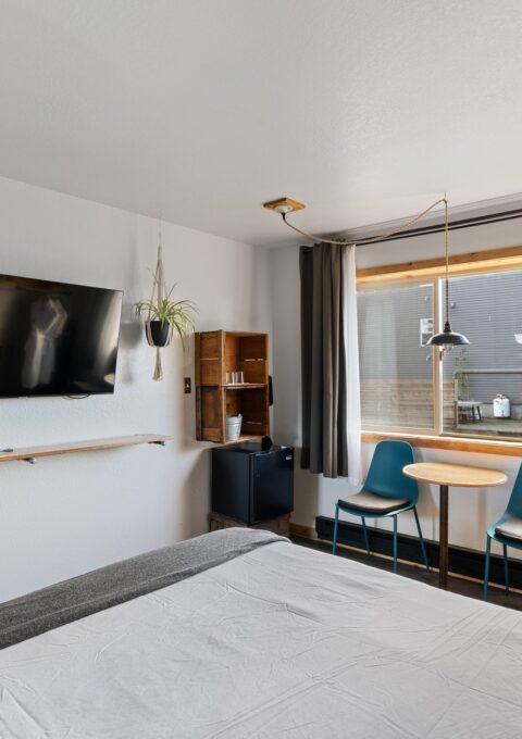 A cozy, modern bedroom features a wall-mounted TV, a small table with two chairs by a window with gray curtains, a bed partially visible in the foreground, and decor including a hanging plant, a framed picture, and a wooden shelf.