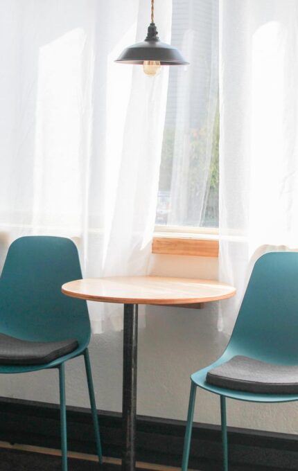 A small, round wooden table with a black metal base is positioned in front of a window with sheer white curtains and blackout drapes. Two teal chairs with gray cushions are placed on either side of the table. A black pendant lamp hangs above.