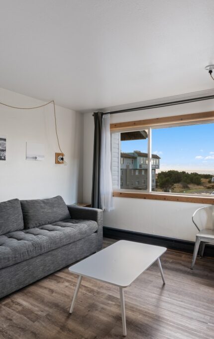 A small living room with a gray sofa, white coffee table, and wicker basket. A window with curtains offers an outdoor view. A wall-mounted plant and framed pictures decorate the white walls. A wooden desk with chairs and hanging light fixtures is on the right side.