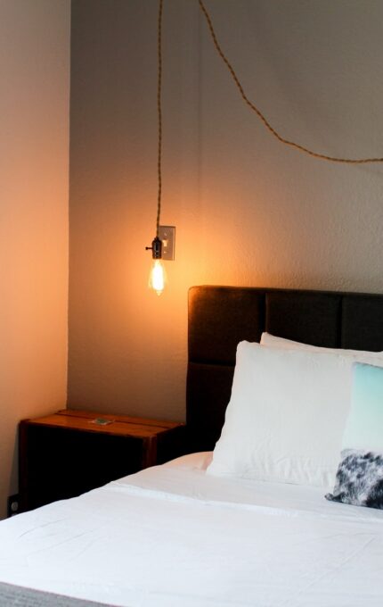 A bedroom with a neatly made bed, featuring white linens and a pillow with a mountain design. A single light bulb hanging from the wall illuminates the room. A wooden nightstand is beside the bed, and there is a window with wooden trim on the left side.