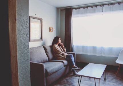 A person with curly hair sits on a gray couch in a living room, looking out the window. The room has wooden floors, a coffee table, and a white chair by the window. A framed picture hangs on the wall above the couch, and the window has dark curtains and white sheer curtains.