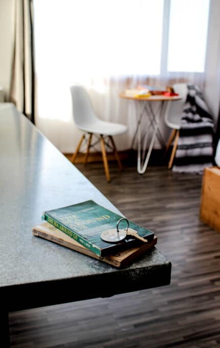 A tabletop displays two stacked books and a pair of glasses. In the background, a small round table with two white chairs and a window with light-colored curtains are visible. The flooring is wooden, and a blanket is draped over one of the chairs.