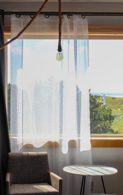 A room with two beige upholstered chairs, a small round table, and a large window with white sheer curtains. The window offers a view of greenery and the ocean in the distance. A hanging light bulb is visible, and a framed black and white photograph is on the wall.