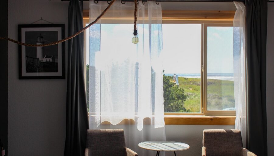 A room with two beige upholstered chairs, a small round table, and a large window with white sheer curtains. The window offers a view of greenery and the ocean in the distance. A hanging light bulb is visible, and a framed black and white photograph is on the wall.