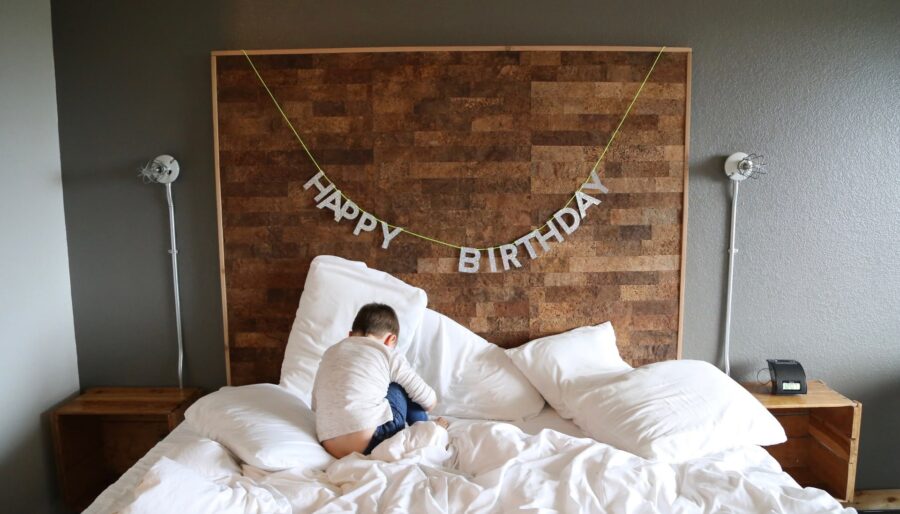 A child wearing a long-sleeved shirt and pants is sitting on a disheveled bed with white linens. A "Happy Birthday" banner is hanging on the headboard above. The room has a minimalist design with wall-mounted lights and small wooden nightstands.
