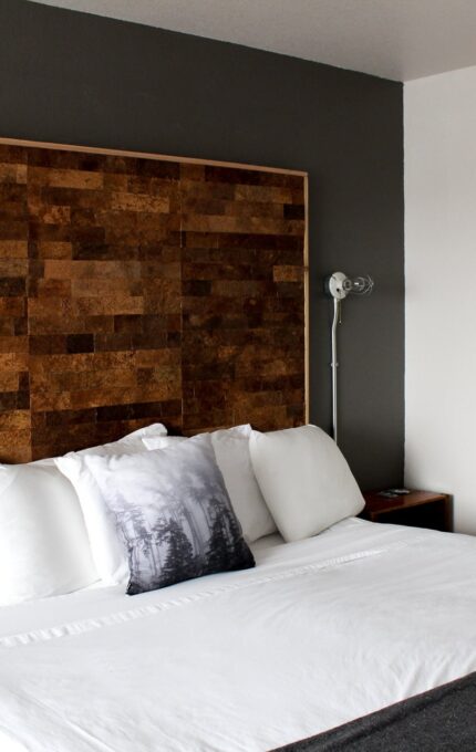 A modern bedroom with a large white bed featuring a wooden headboard. The bed has multiple pillows, including one with a forest design. Two reading lamps are mounted on the wall above the nightstands. A framed black-and-white landscape photograph is hung on the wall.