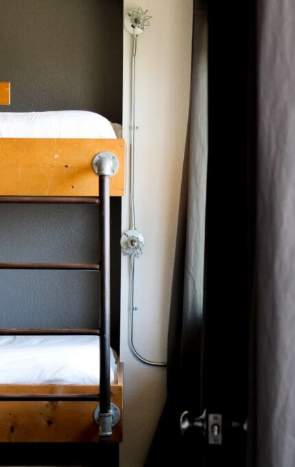 A room with a wooden bunk bed, featuring metal frame details and white bedding. The lower bunk is positioned against a dark wall, and sunlight is streaming in from a window partially covered by a curtain. Electrical conduits are visible on the wall.