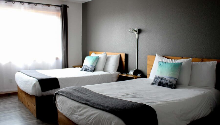 A hotel room with two neatly made double beds. Each bed has white bedding, a grey throw blanket at the foot, and a pillow with a blue and white design. The room has a grey accent wall, a bedside table with a lamp, and a window with white curtains on the left.