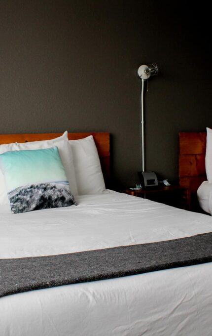 A hotel room with two made-up double beds placed side by side against a dark gray wall. Each bed has a wooden headboard, white linens, and a decorative pillow with a light blue design. There is a wall-mounted reading lamp and a bedside table between the beds.