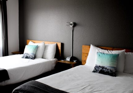 The image shows a hotel room with two double beds. Each bed has white linens and two pillows. The beds are positioned against a dark gray wall with a bedside table in between. A lamp and a landline phone are on the bedside table. The room has natural light.