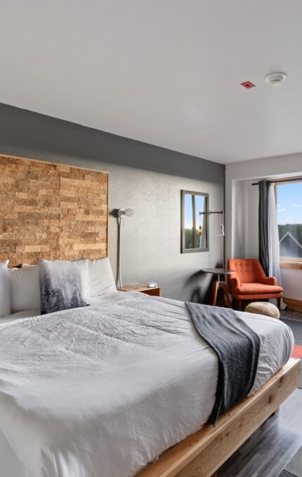 A modern hotel room with a large bed in the center, featuring white linens and a dark gray blanket. The room has a natural wood headboard and nightstands, a large window with a view of trees, an orange armchair, and a small black refrigerator.