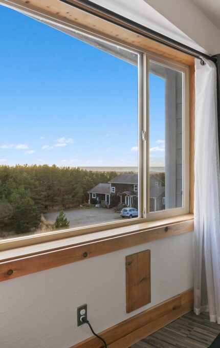 A room with a large window overlooking a house surrounded by trees. The room has a chair, a small round table, artwork with colorful geometric designs on the wall, and curtains on the window. The sky outside is clear and blue.