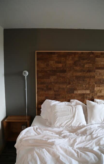 A neatly made bed with white linens and pillows is positioned against a wall with a wood-textured headboard. To the left of the bed, there is a simple wooden nightstand and a wall-mounted reading lamp. The room has a minimalist design.