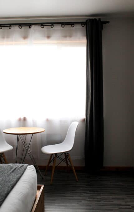 A minimalist room with a round wooden table and two white chairs in front of a window with white curtains and dark drapes. The corner of a bed with gray bedding is visible in the foreground. The floor has dark laminate wood flooring.