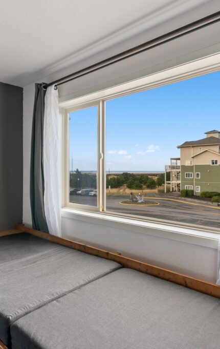 A sunlit room with a large window features a simple, minimalist bed with grey bedding. Three black-and-white photos hang on the grey wall next to the bed. Outside the window, a building and a winding road are visible under a clear blue sky.