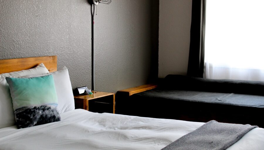 A hotel room features a neatly made bed with white linens, a gradient turquoise and white pillow, and a fuzzy gray pillow. Next to the bed is a wall lamp and a small wooden side table. A dark cushioned bench is placed by the window with white curtains.