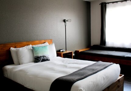 A bedroom with a large bed featuring white linens and a gray blanket. A wooden bed frame and nightstand complement the decor. A floor lamp stands next to the bed, and a window with sheer curtains allows natural light to enter the room. There is a daybed beside the window.