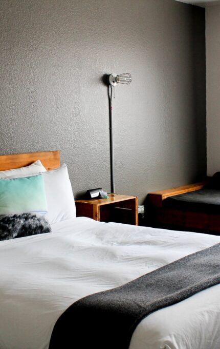 A bedroom with a large bed featuring white linens and a gray blanket. A wooden bed frame and nightstand complement the decor. A floor lamp stands next to the bed, and a window with sheer curtains allows natural light to enter the room. There is a daybed beside the window.