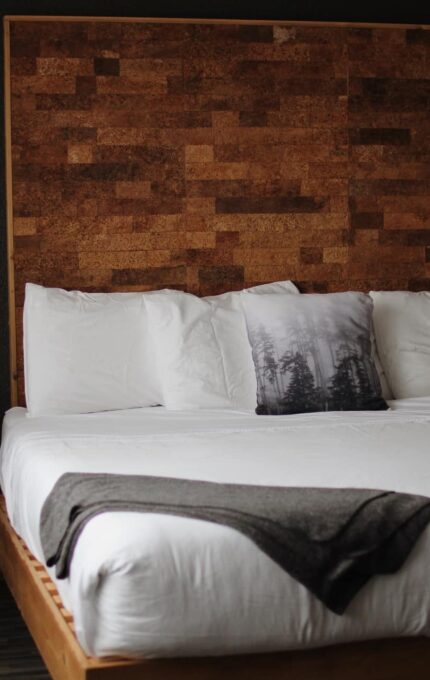 A neatly made bed with white linens, featuring a large wooden headboard with a geometric pattern. There are two white pillows and one decorative pillow with a forest design. A gray blanket is draped over the foot of the bed. Two wall-mounted lamps are on either side of the headboard.