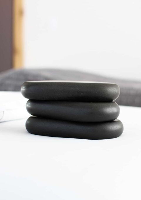 Three black stones are stacked on top of each other on a white surface. The background includes a gray blanket and a door frame. The overall setting appears calm and minimalistic.