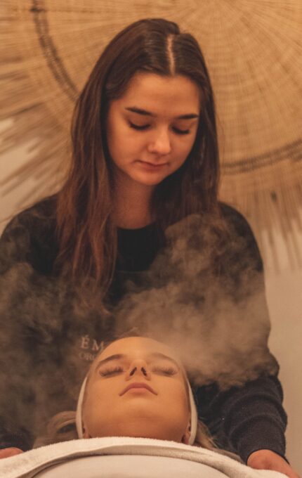 A woman with long hair is providing a facial treatment to another woman lying down with eyes closed. There is steam around the face of the woman receiving the treatment. A woven circular decor piece hangs on the wall in the background.