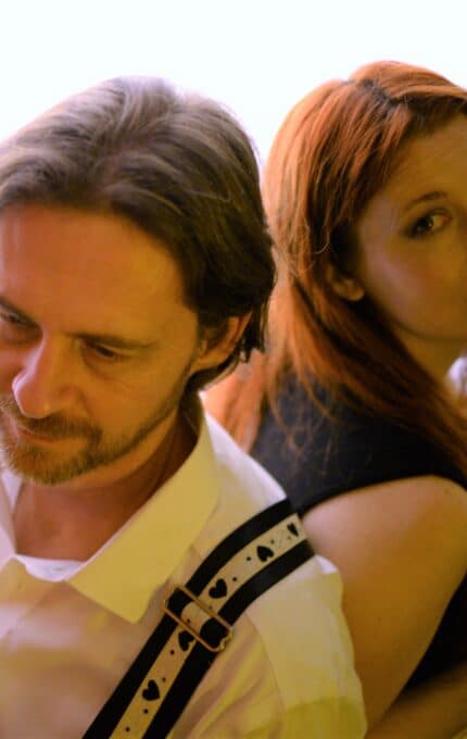 A man with a beard and a woman with red hair are seated back-to-back. The man is wearing a white shirt and holding a banjo, while the woman is in a black top, looking at the camera. The background is softly lit.