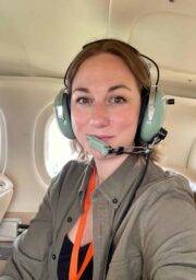 A person with shoulder-length light brown hair wearing a black shirt, olive green jacket, and orange lanyard is sitting in an aircraft while wearing a headset. The person is smiling and looking at the camera. Beige seats and windows are visible in the background.