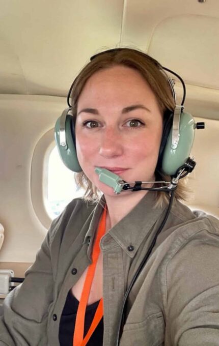 A person wearing a green shirt, black top, orange lanyard, and green aviation headset is sitting inside an aircraft. The person appears to be facing the camera, with aircraft windows visible in the background.