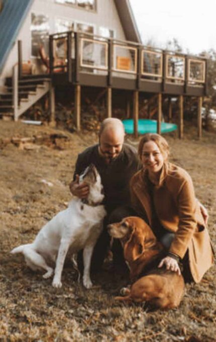 A couple kneels on the grass with two dogs outside a house with a steep, blue roof. The man, wearing a dark jacket, pets one dog, while the woman in a tan coat sits beside them, smiling. The house has a raised deck and the yard is expansive with trees in the background.