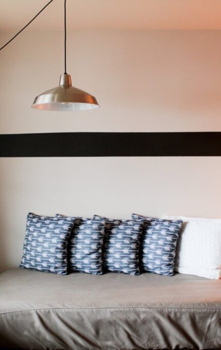 A small room features a single bed against a wall with five patterned blue pillows and one white pillow. Above the bed, a metal pendant light hangs from the ceiling. A window with closed curtains is on the left, and a lit lamp and clock sit on a wooden side table.