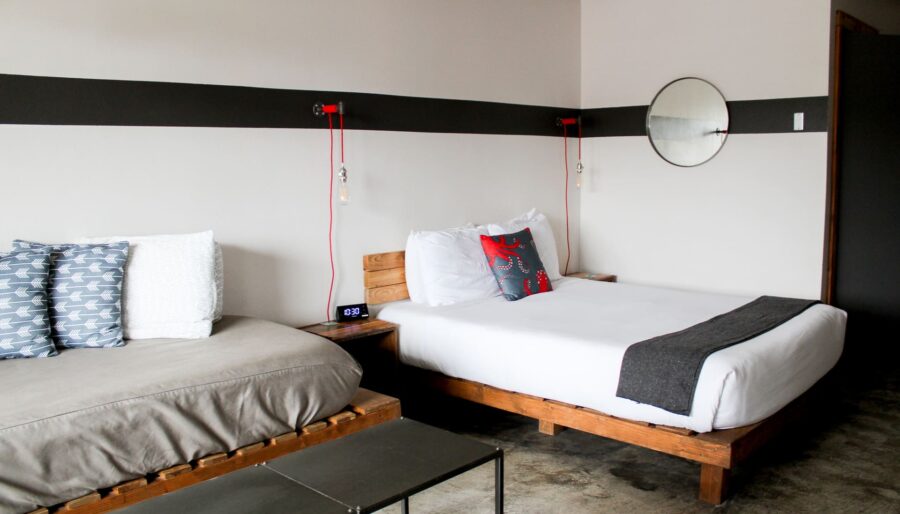 A modern hotel room featuring a bed with white linens, decorative pillows, and a gray throw blanket. A sofa with cushions is positioned to the left. The room has minimalistic decor including a wall-mounted round mirror and hanging bedside lamps.