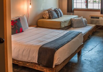 A small bedroom with a wooden platform bed, white bedding, and a gray blanket. The room has an additional daybed with multiple cushions, a hanging light bulb, and an air conditioning unit below the window. The floor is polished concrete.