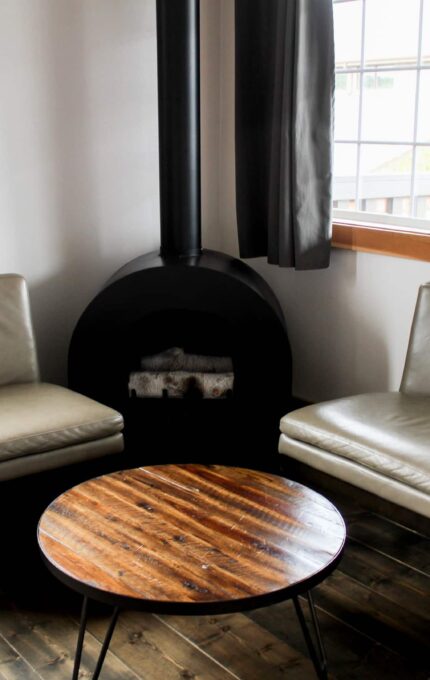 A small seating area with two gray leather chairs positioned around a circular wooden coffee table. Behind the chairs is a black cylindrical stove, and large windows with gray curtains provide natural light to the space.