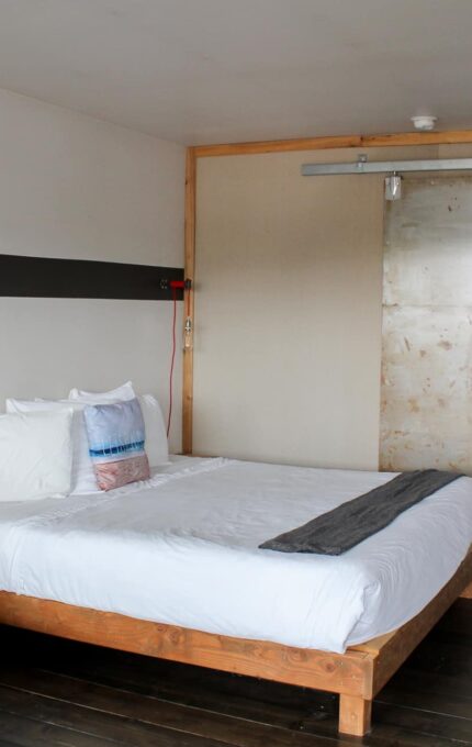 A modern hotel room features a queen-sized bed with white linens and several pillows. The room includes a gray chair, a sliding rustic metal door, and a desk with a wooden surface. The flooring is dark wood, and the walls are light-colored.