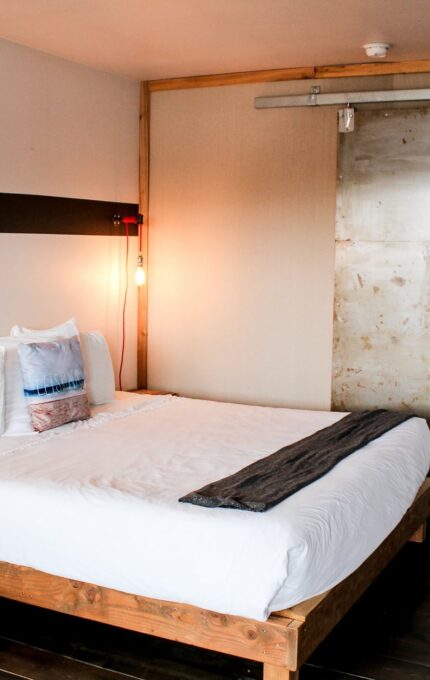 A minimalist hotel room with a queen-sized bed featuring white linens and multiple pillows. The room has a wooden floor, a leather chair, two exposed lightbulbs as bedside lamps, and a sliding barn door leading to another area. A desk and chair are visible on the right.