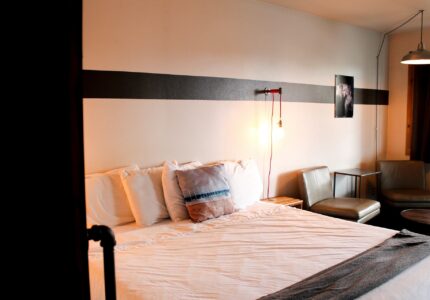 A neatly made bed with white linens and several pillows is positioned against a white wall with a horizontal dark stripe. The room features two chairs, a small table, a wall-mounted light, and framed artwork. The atmosphere is cozy and minimalist.